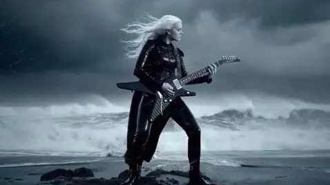 Woman with long blonde hair playing electric guitar on a rocky shore with crashing waves and a stormy sky.
