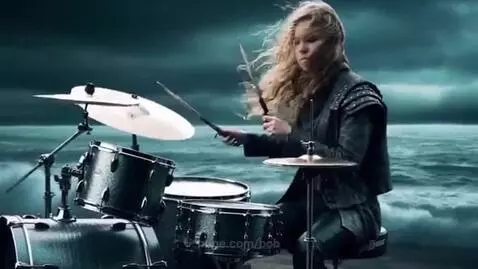 Woman with long blonde hair playing drums on a stormy beach with large waves and wind.