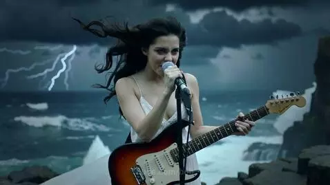 Woman with long dark hair playing electric guitar and singing on a rocky cliff by a stormy ocean.