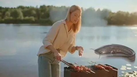 Woman grilling shashlik by a river on a sunny day, with a catfish visible in the water.