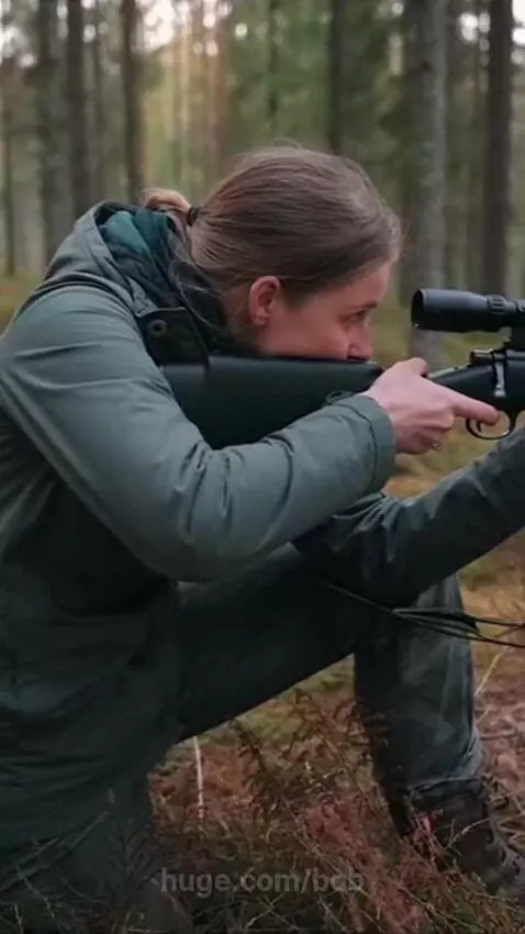 Woman in green jacket aiming a rifle with scope at a wild boar in a forest.