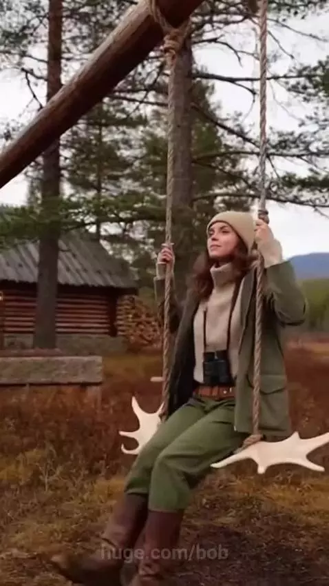 Woman in outdoor attire swinging on a wooden swing with a seat made of moose antlers in a taiga forest.