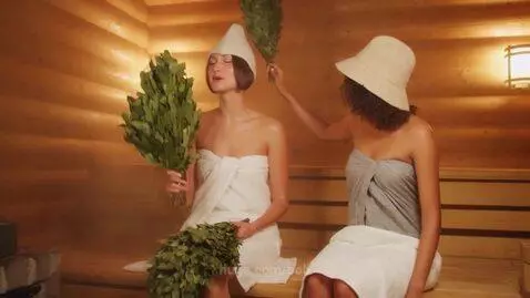 Two women in towels and sauna hats relax in a steamy wooden sauna, one holding a bundle of leaves.
