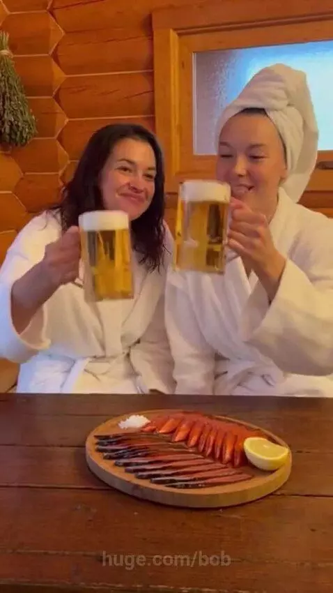Two women in bathrobes smile and clink beer mugs at a table with dried fish after a sauna.