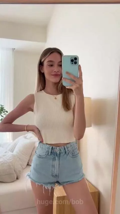 Young woman with long blonde hair in a cream crop top and denim shorts takes a mirror selfie, smiling.