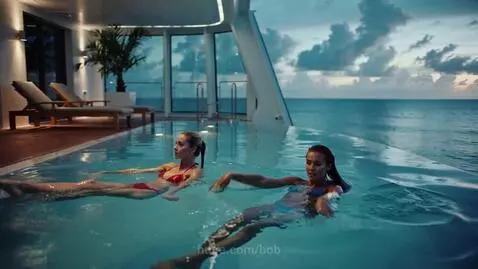 Two women in bikinis swimming and splashing in an infinity pool with a vast ocean view.