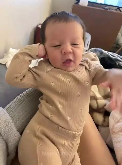 Close-up of an adorable baby with dark hair in a light brown onesie, making cute hand gestures and facial expressions while held upright.