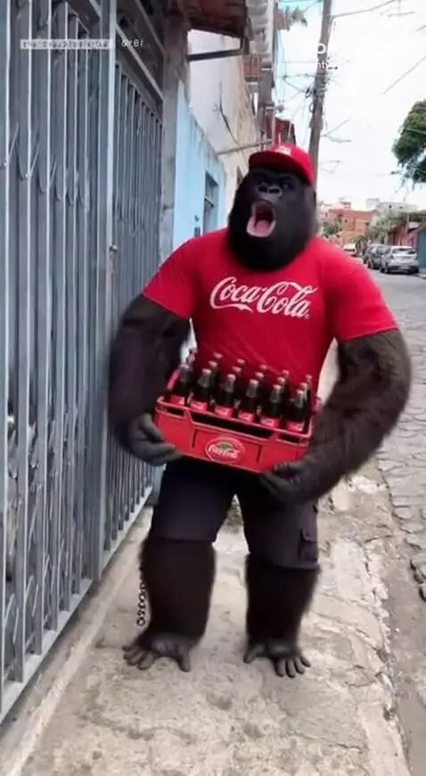 Person in a gorilla costume wearing a Coca-Cola shirt, holding a crate of Coca-Cola bottles on a street.