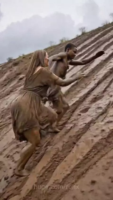 Man and woman covered in mud, laughing as they slide down a muddy hill.