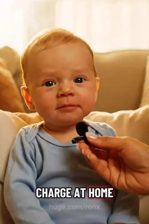 Baby in a blue onesie on a couch, looking at the camera while a microphone is held near its mouth.