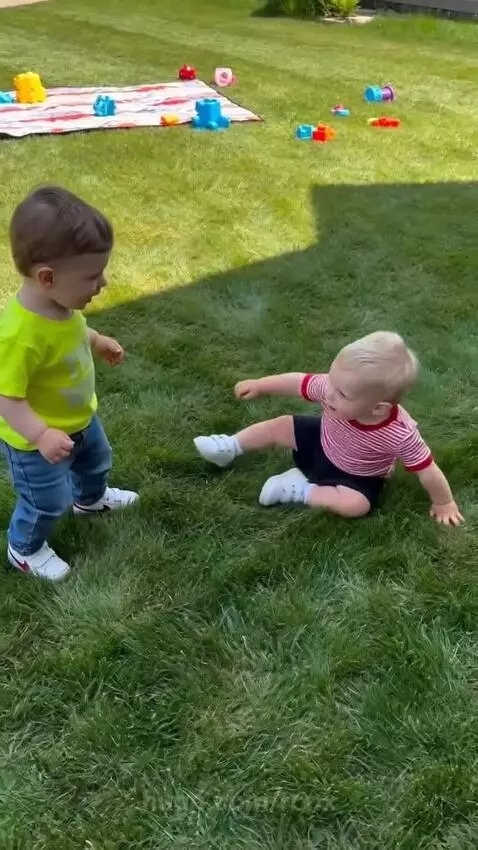 Two toddlers playfully wrestling and laughing on a green lawn with toys and a blanket.