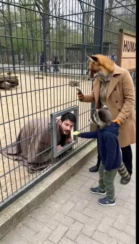 Man in a cage being fed by a child wearing a raccoon mask.