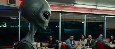 Close-up of a grey alien with large black eyes in a diner, patrons look terrified.