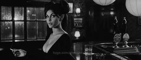 Man in trench coat and woman in low-cut dress in a dimly lit bar, engaged in suggestive conversation.