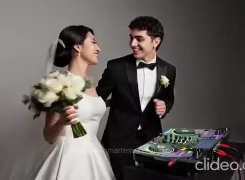 Bride in white gown and groom in tuxedo smiling behind a DJ mixer, ready to play music.