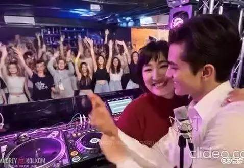 Couple at DJ booth smiling and laughing, with a crowd cheering in the background at a club.