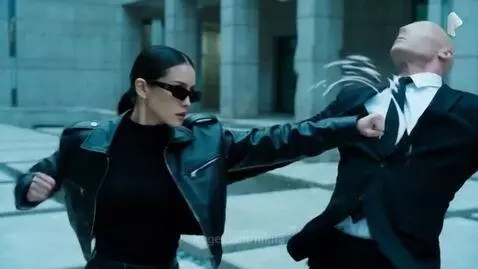 Woman in black leather jacket and sunglasses performing martial arts combat against men in suits.