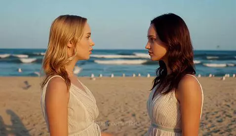 Two young women in dresses on a beach, arms around each other, looking surprised.