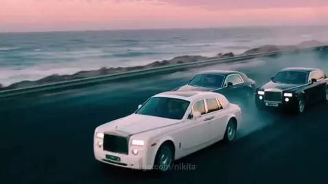 Three Rolls-Royce Phantom cars, white and black, driving on a coastal road with smoky trails at sunset.