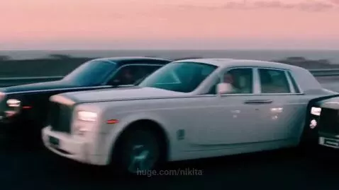 Three Rolls-Royce Phantom cars, white and black, driving on a coastal road with smoky trails at sunset.