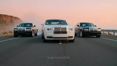 Three Rolls-Royce Phantom cars, one white and two black, driving on a scenic coastal highway at dusk or dawn, with one performing a smoky drift.