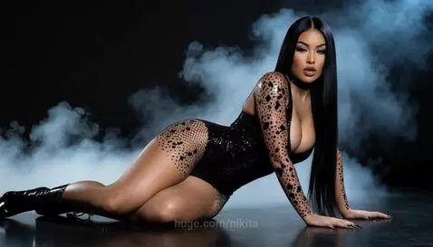 Woman with long dark hair in a black sequined bodysuit and high heels, posing suggestively on a dark reflective surface with smoke.