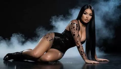 Woman with long dark hair in a black sequined bodysuit and high heels, posing suggestively on a dark reflective surface with smoke.