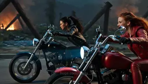 Two women in leather motorcycle gear with glowing orange eyes race powerful Harley-Davidsons down a burning, desolate street.