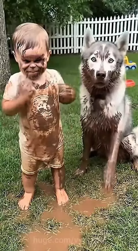 A young child and a husky dog, both completely covered in mud, stand in a muddy puddle on a lawn.