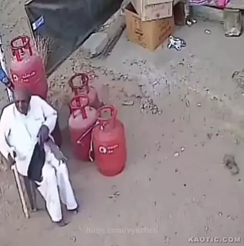 Elderly man in white outfit on chair is hit by a bull, falling to the ground with gas cylinders.