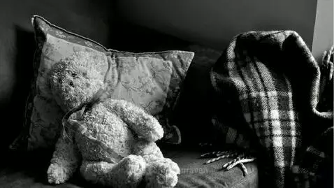 Black and white image of a teddy bear on a couch, with a plaid blanket.