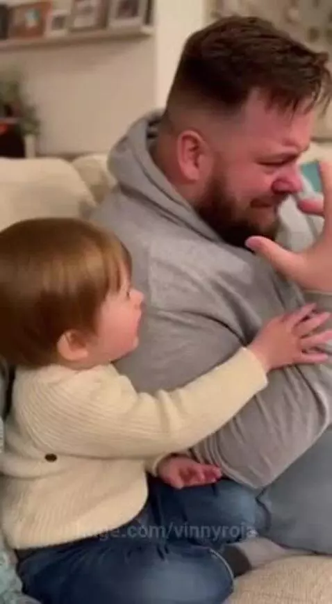 Bearded man in grey hoodie laughs as a child in a cream sweater hugs him.