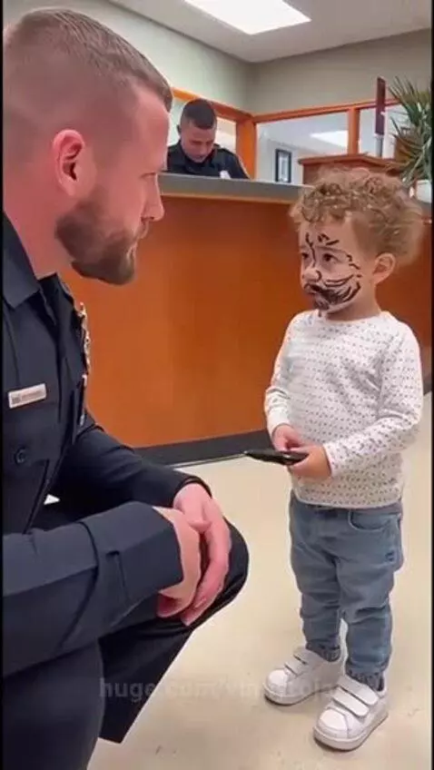 Child with drawn-on beard and eyebrows kneels and talks to a police officer at a station.