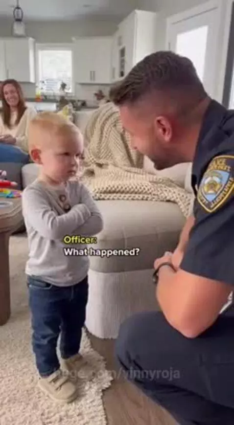 A serious toddler with arms crossed talks to a kneeling police officer about his mom eating his chocolate.