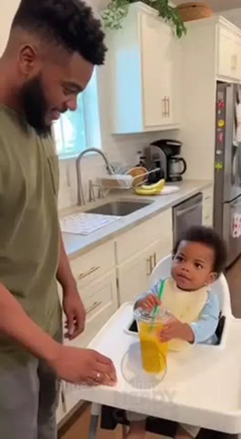 Bearded dad in kitchen smiling at baby in high chair, handing baby a drink.