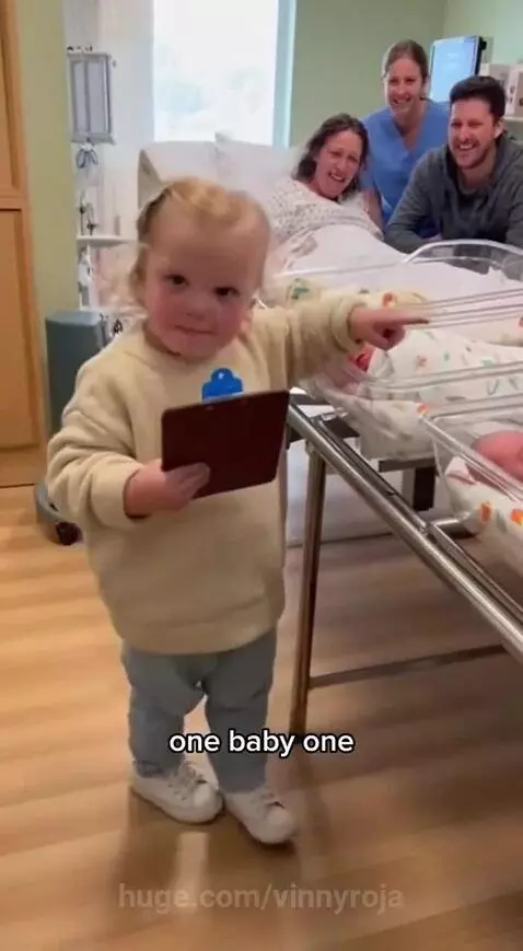 Toddler with clipboard pointing at three newborn babies in a bassinet, reacting with surprise.