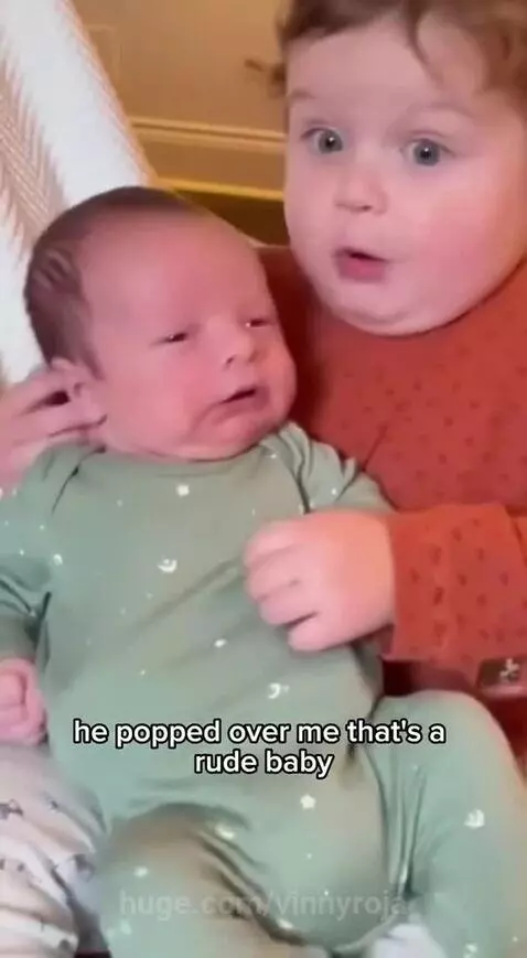 Toddler with wide eyes looking at a newborn baby on a couch, reacting to a sound.