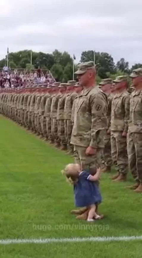 Soldier in uniform embracing a young child in a blue dress on a grassy field.