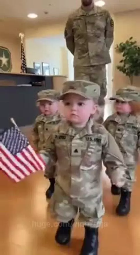 Three toddlers in camouflage uniforms and hats march in a line, one holding an American flag, with soldiers in the background.