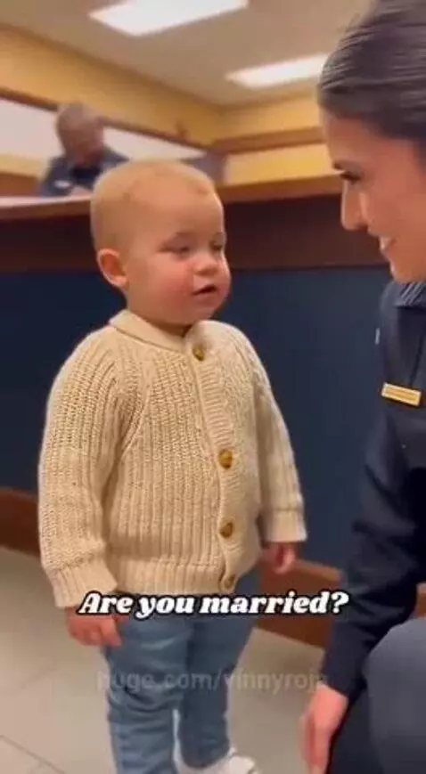 A toddler in a cream cardigan asks a crouching police officer if she is married.