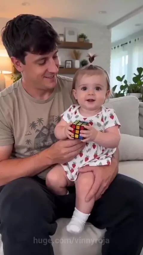 Father holding a baby girl in a strawberry dress, who is holding a Rubik's cube.