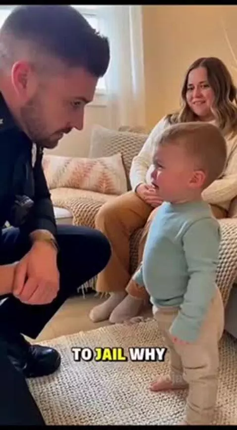 A toddler cries while talking to a police officer, with a pregnant woman smiling in the background.