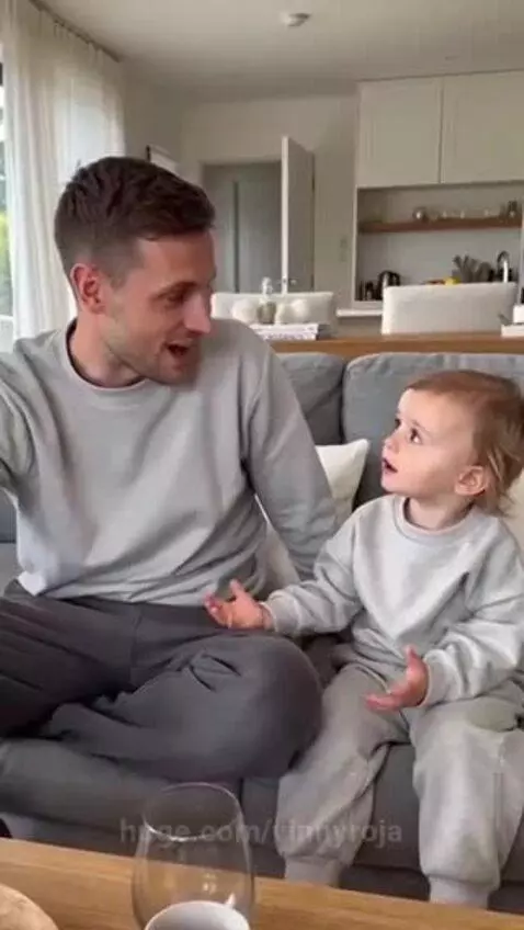 Father and child in matching grey outfits sitting on a couch, child looking at father with a curious expression.
