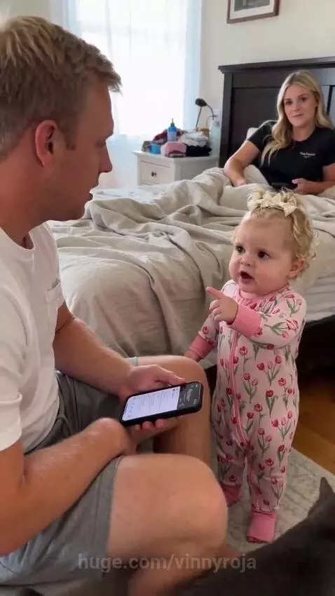 Baby girl in a pink floral onesie points at her dad, who is on his phone, asking about kisses.