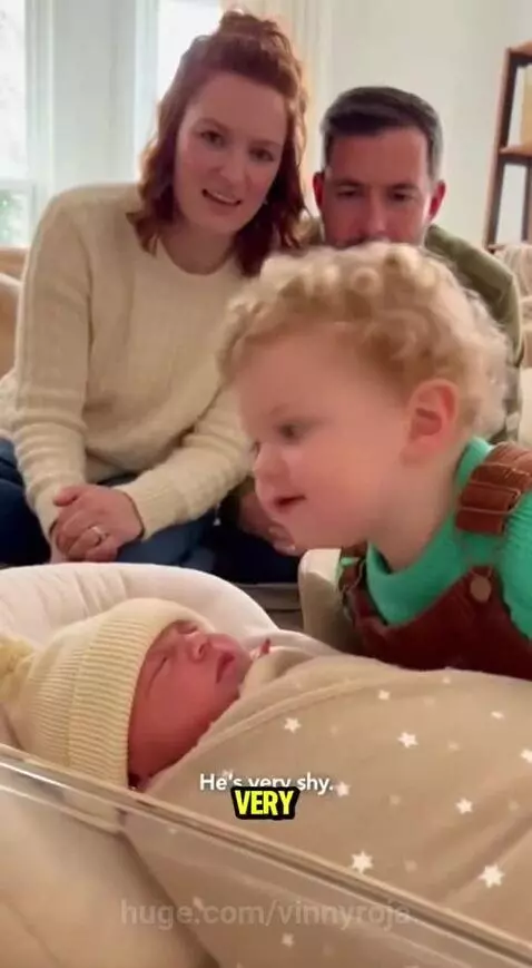 Toddler leaning over a bassinet, talking to a newborn baby. Parents watch and laugh in the background.