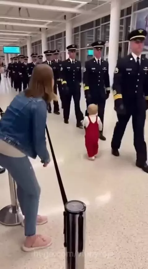 Pilot in uniform kneeling to hug a baby dressed in red overalls in an airport terminal.