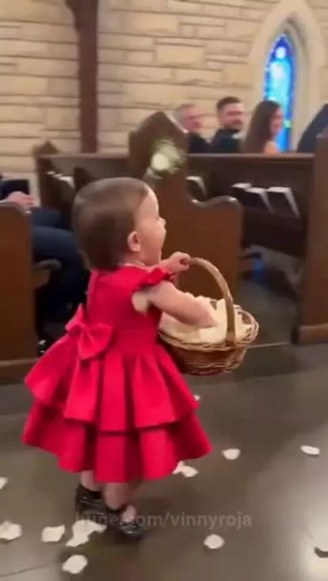 Young flower girl in a red dress scattering a large handful of white petals down a church aisle.