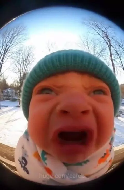 Close-up fisheye view of a crying baby wearing a teal hat and patterned blanket, with a snowy background.