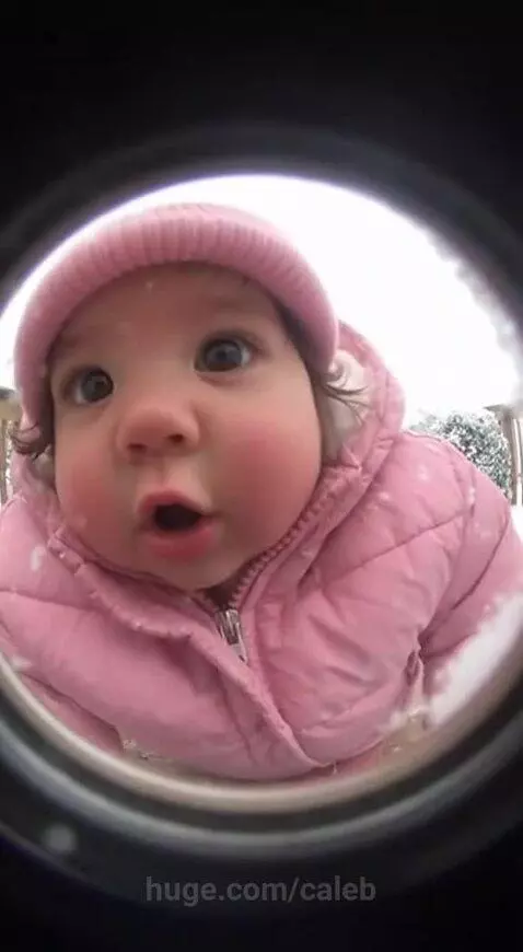 A young child in a pink winter coat and hat smiles joyfully through a peephole, snowflakes on their face.