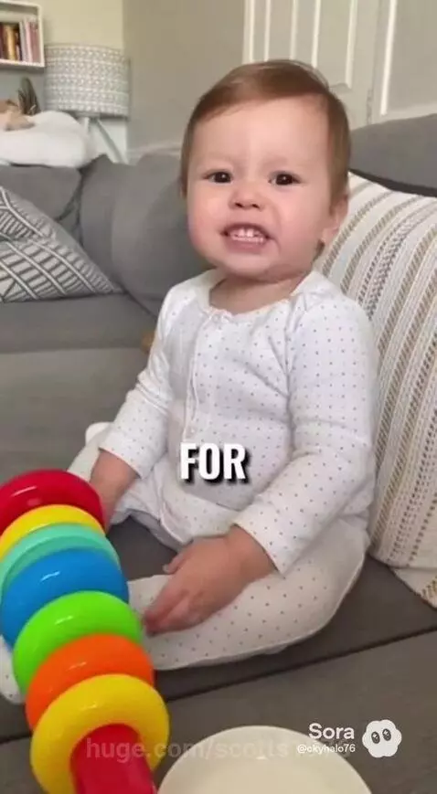 Baby with light brown hair in a polka-dotted onesie sitting on a couch, looking at the camera and seemingly asking for a sandwich.